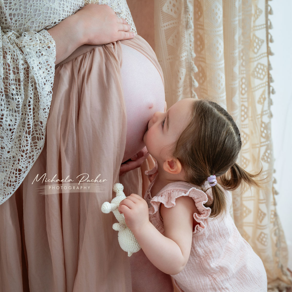 Kleines Mädchen küsst den Babybauch ihrer schwangeren Mama, während sie ein gehäkeltes Stofftier in der Hand hält; beide tragen zarte Rosé- und Cremetöne.